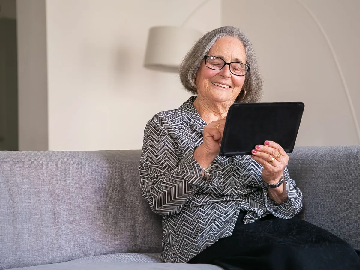 Oudere vrouw met bril zit glimlachend op de bank en gebruikt een tablet