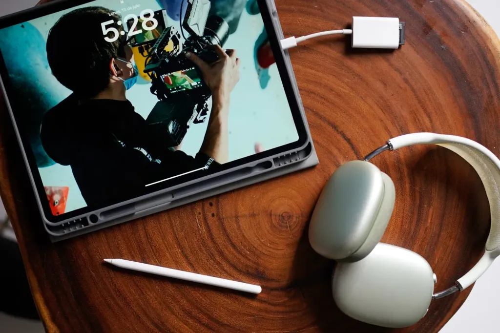 iPad met aangesloten oplaadkabel op een houten tafel, naast een koptelefoon en een Apple Pencil