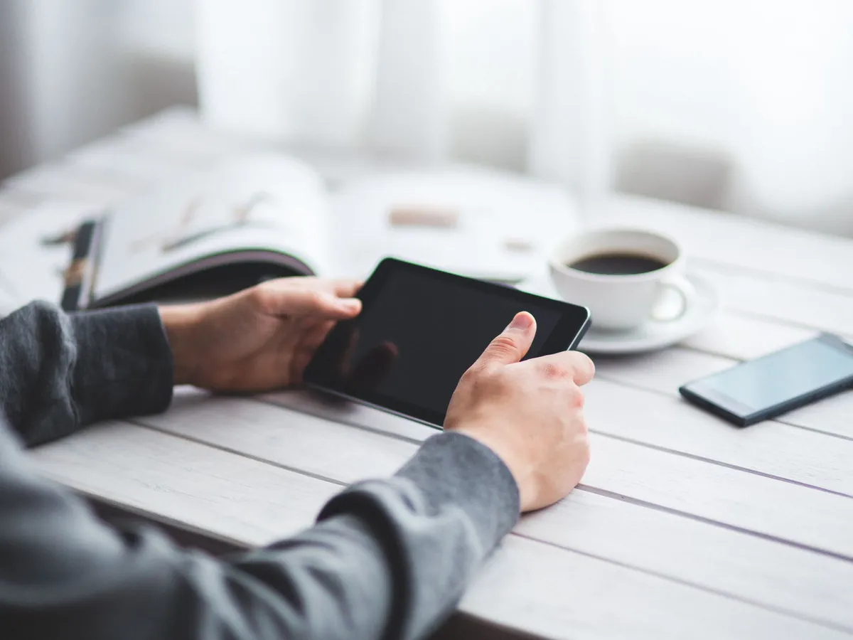 Iemand houdt een kleine zwarte tablet vast aan een lichte tafel met koffie en een smartphone