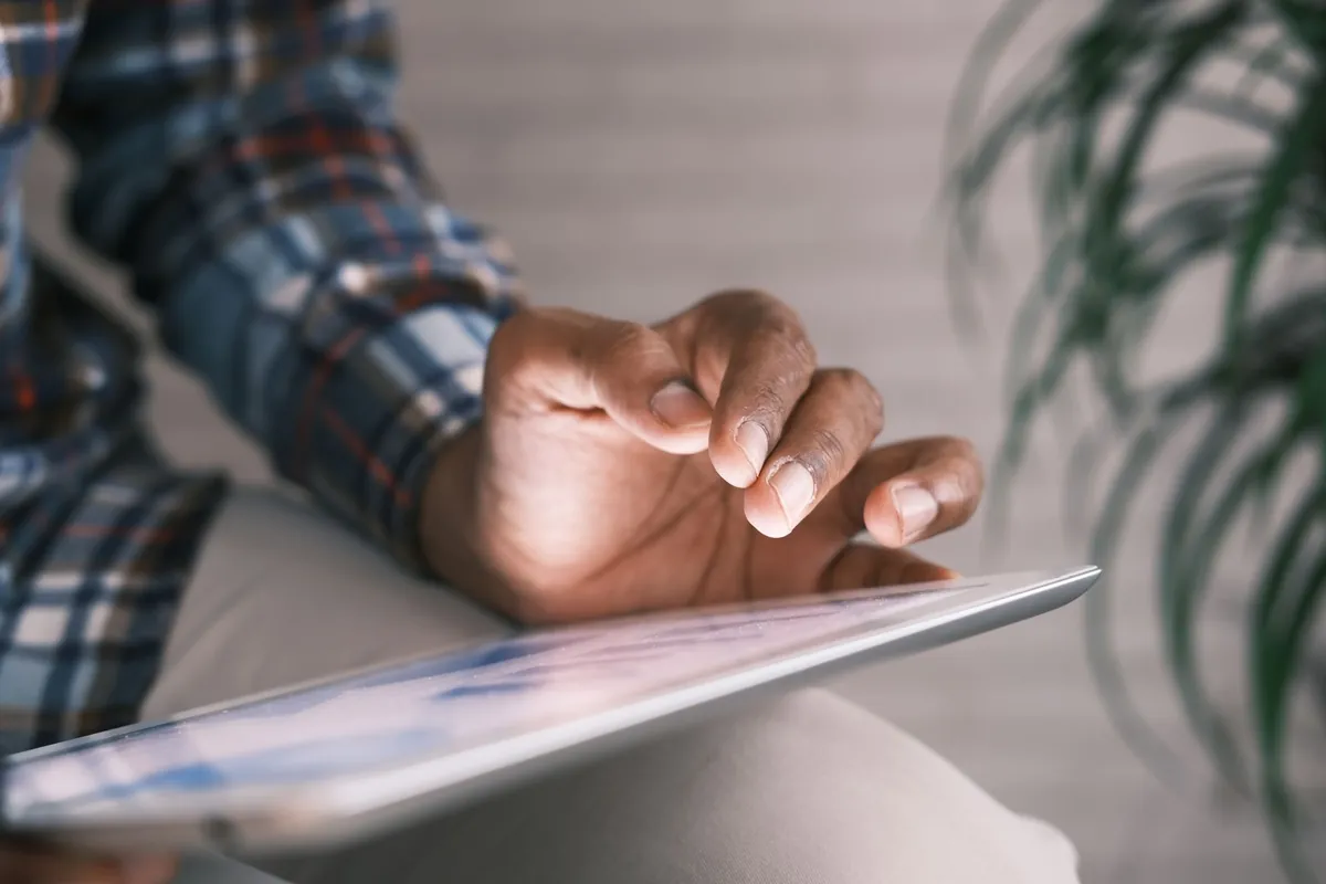 Hand boven het scherm van een tablet op schoot, binnen bij een plant